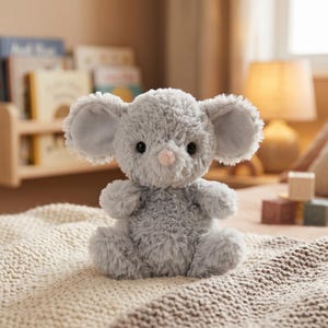 Peut inclure: Une peluche grise en forme de souris avec de grandes oreilles, des yeux noirs et un nez rose. L'animal en peluche est assis sur une couverture en tricot cr&egrave;me. Le jouet est con&ccedil;u pour les enfants.