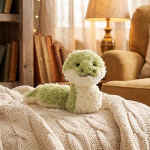 May include: A plush snake toy with a green head and body, and a white belly. The snake is lying on a cream-colored cable-knit blanket. The background includes a bookshelf, a lamp, and a cozy armchair.