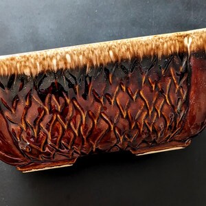 May include: A rectangular ceramic planter with a brown and tan glaze. The planter has a textured diamond pattern on the sides and a glossy finish. The top edge is a lighter tan colour, and the interior is dark brown.
