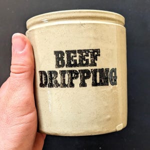 May include: A beige ceramic jar with the words "BEEF DRIPPING" printed in black, block letters. The jar has a cylindrical shape with a slightly flared rim. The jar is held in a hand.
