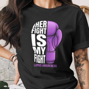 May include: Black t-shirt with a purple boxing glove graphic. The text reads "HER FIGHT IS MY FIGHT" in white, with "LUPUS AWARENESS" in purple below. A design promoting awareness.