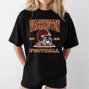May include: Black t-shirt with a distressed graphic. The design features the text "WASHINGTON FOOTBALL", a football helmet, and the text "EST. 1932". The shirt has a crew neck and a football field graphic. The shirt is black.