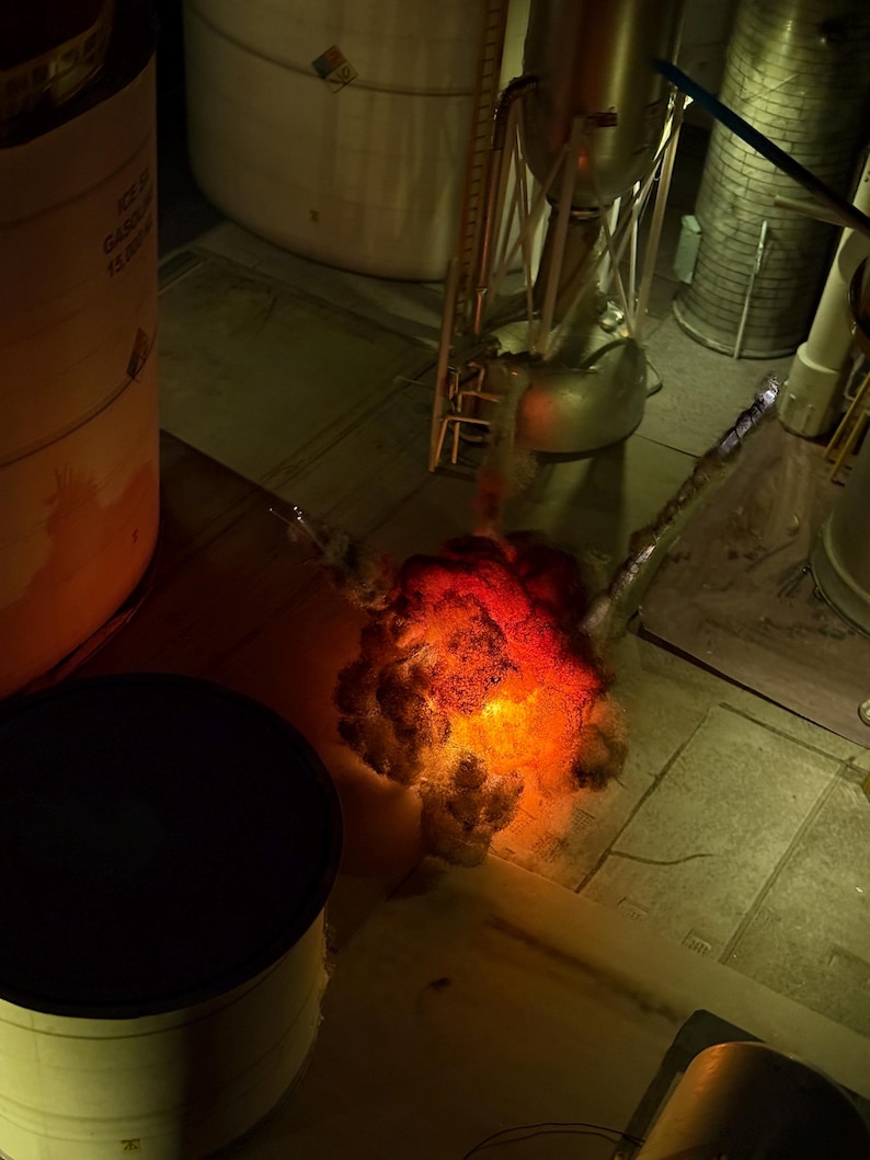 Pu&ograve; includere: Un diorama in miniatura che mostra una scena di lancio di un razzo con un effetto di esplosione arancione e rosso brillante alla base. Il modello include una struttura metallica del razzo, serbatoi cilindrici e componenti industriali.