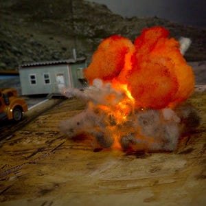 Pu&ograve; includere: Un diorama in miniatura che presenta un effetto esplosione di colore arancione e giallo brillante realizzato in materiale fibroso, posizionato accanto a un piccolo edificio modello e a un camion giocattolo giallo su una superficie di legno.