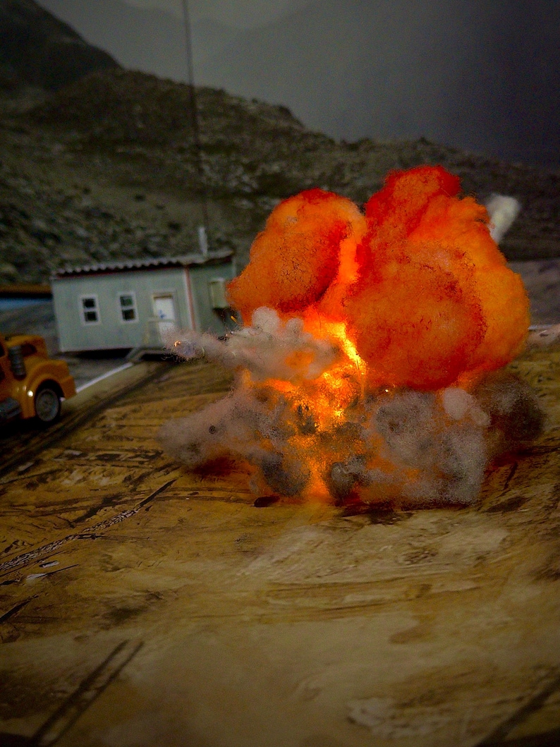 Pu&ograve; includere: Un diorama in miniatura che presenta un effetto esplosione di colore arancione e giallo brillante realizzato in materiale fibroso, posizionato accanto a un piccolo edificio modello e a un camion giocattolo giallo su una superficie di legno.