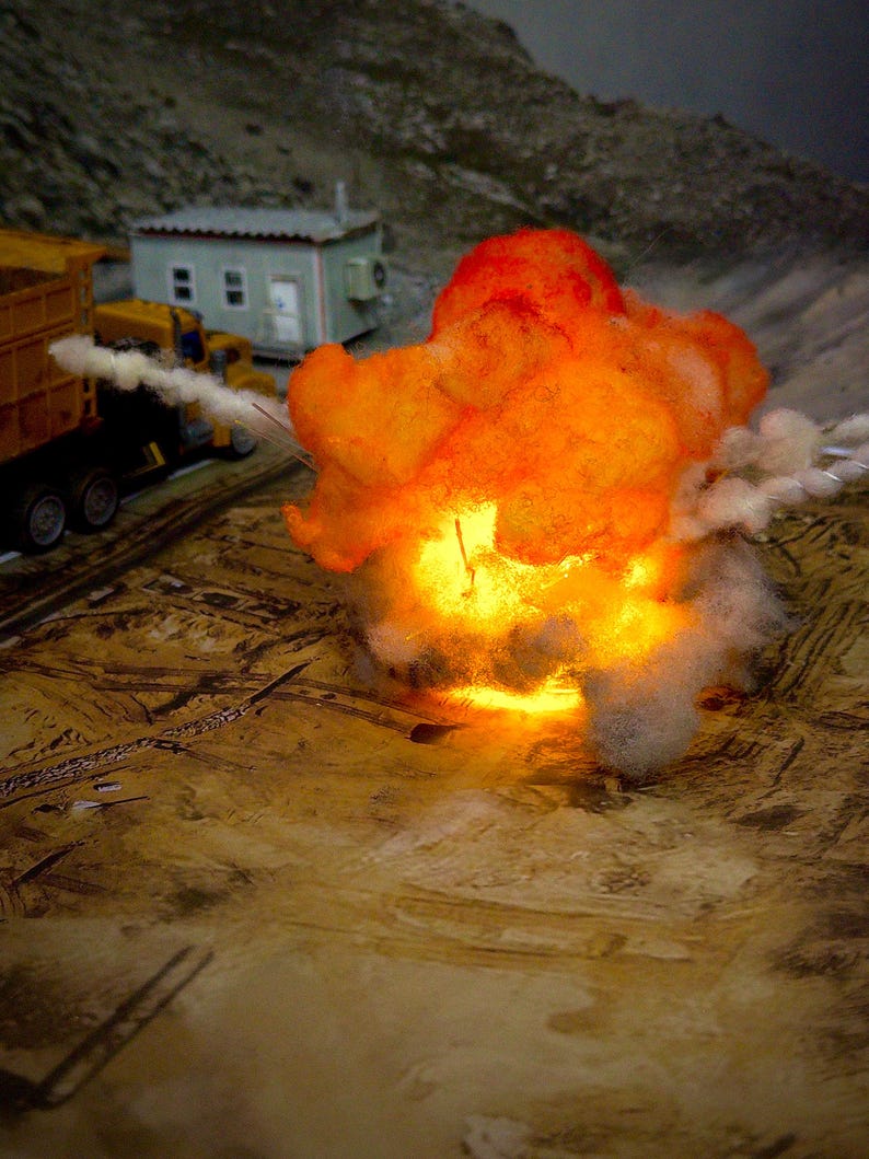 Pu&ograve; includere: Un diorama in miniatura con un effetto esplosione in fibra ottica color arancione e giallo brillante. A sinistra sono visibili un piccolo camion con cassone ribaltabile giallo e un edificio rettangolare chiaro su una superficie marrone.