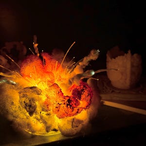 Pu&ograve; includere: Una scultura luminosa con una base strutturata simile a una nuvola in tonalit&agrave; grigio scuro e nero, illuminata dall'interno da una luce arancione e gialla brillante. Sottili fili di fibra ottica traslucidi si estendono verso l'esterno.