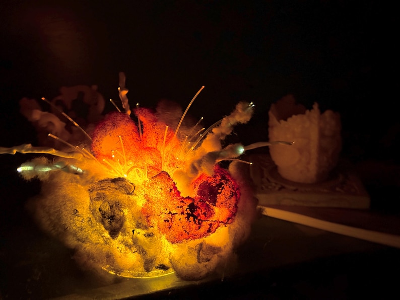 Pu&ograve; includere: Una scultura luminosa con una base strutturata simile a una nuvola in tonalit&agrave; grigio scuro e nero, illuminata dall'interno da una luce arancione e gialla brillante. Sottili fili di fibra ottica traslucidi si estendono verso l'esterno.