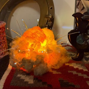 Pu&ograve; includere: Una scultura luminosa a fibre ottiche a forma di esplosione, caratterizzata da un nucleo strutturato simile a una nuvola nei toni dell'arancione e del giallo. Sottili fili di fibra ottica traslucidi si estendono verso l'esterno, emettendo piccoli punti luce.