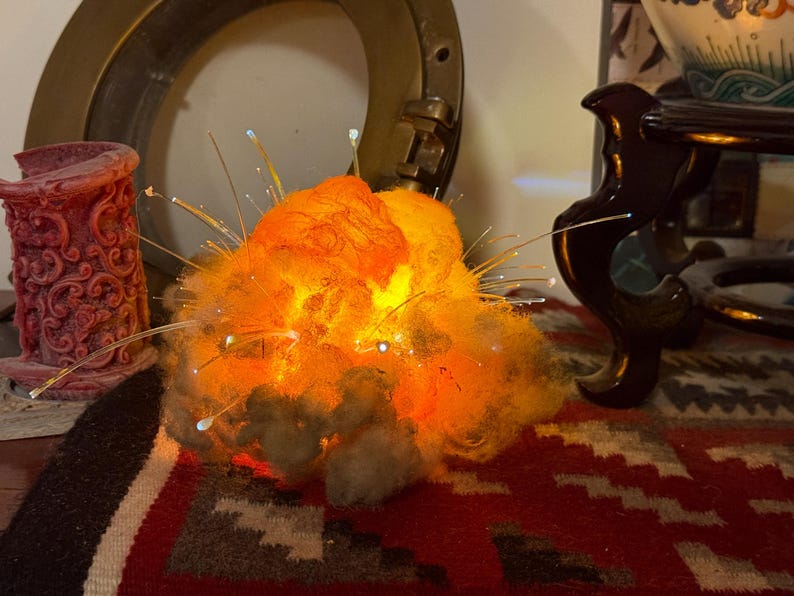 Pu&ograve; includere: Una scultura luminosa a fibre ottiche a forma di esplosione, caratterizzata da un nucleo strutturato simile a una nuvola nei toni dell'arancione e del giallo. Sottili fili di fibra ottica traslucidi si estendono verso l'esterno, emettendo piccoli punti luce.