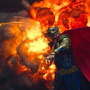 Pu&ograve; includere: Un action figure dettagliato di un guerriero che indossa un elmo alato dorato, un'armatura blu e oro e un mantello rosso fluente, posizionato davanti a una grande esplosione arancione e gialla con fumo scuro denso e scie luminose.
