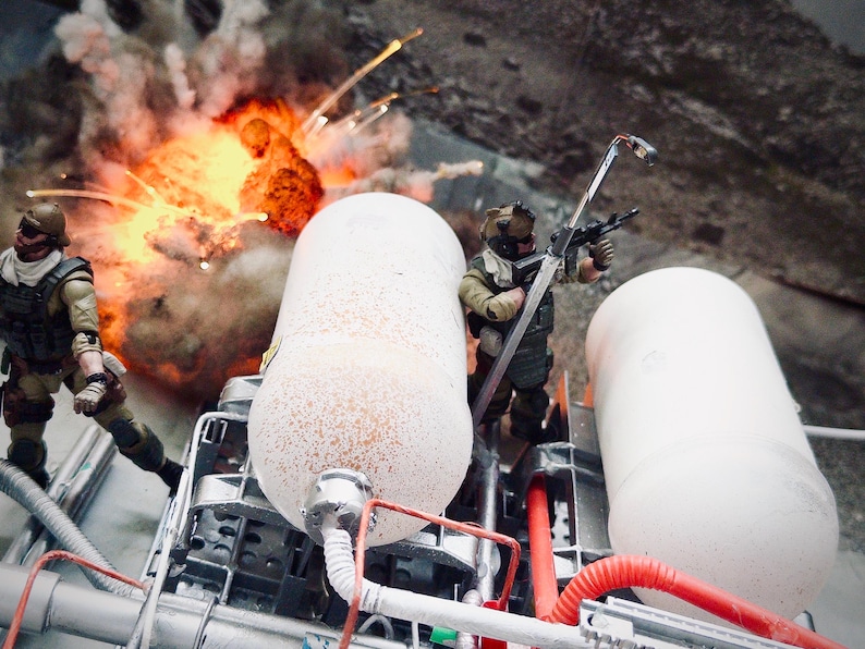 Pu&ograve; includere: Un diorama con due action figure di soldati in miniatura con equipaggiamento tattico posizionate vicino a due grandi serbatoi cilindrici bianchi. Un soldato impugna un fucile mentre sullo sfondo si vede un effetto di esplosione arancione.