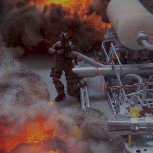 Pu&ograve; includere: Una action figure in miniatura con equipaggiamento tattico si trova vicino a una complessa struttura industriale con tubi e un grande serbatoio bianco. La scena &egrave; circondata da denso fumo grigio e vivaci fiamme arancioni, creando un effetto drammatico.