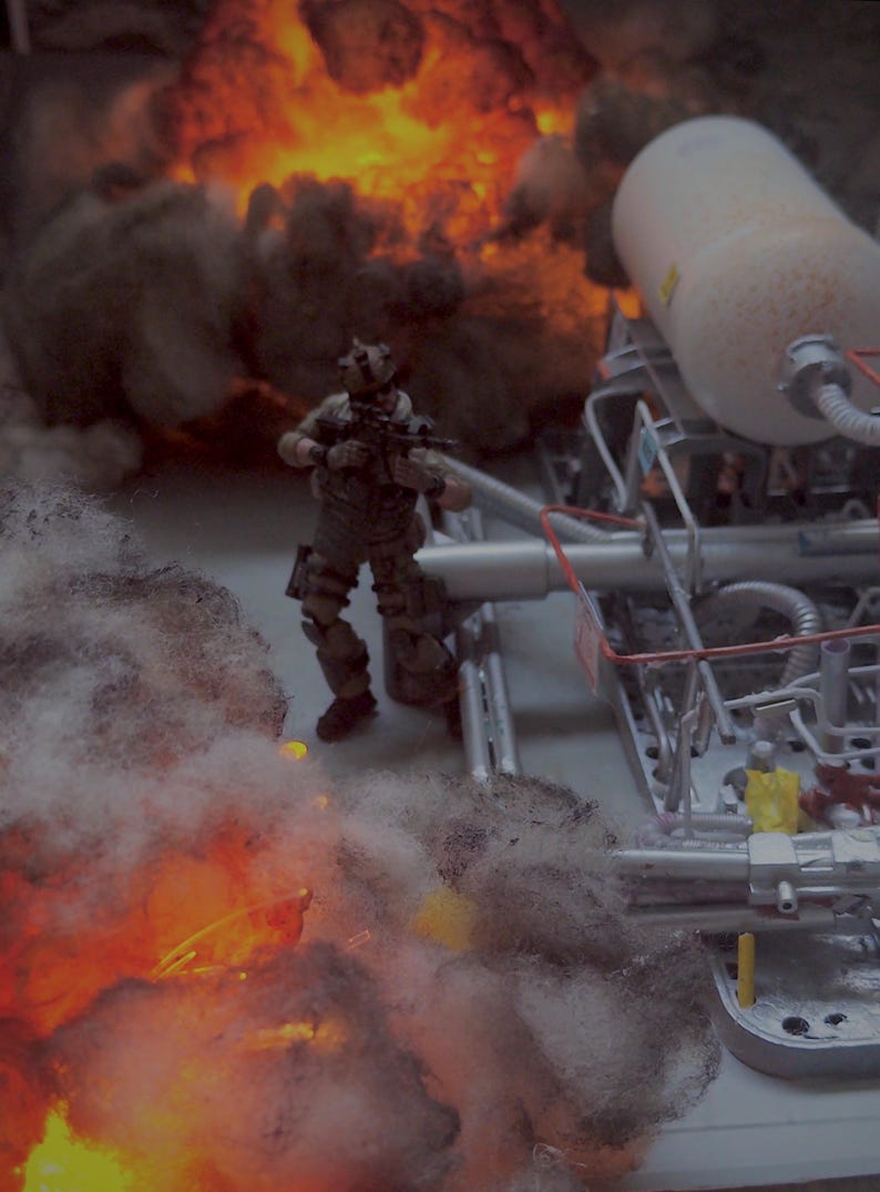 Pu&ograve; includere: Una action figure in miniatura con equipaggiamento tattico si trova vicino a una complessa struttura industriale con tubi e un grande serbatoio bianco. La scena &egrave; circondata da denso fumo grigio e vivaci fiamme arancioni, creando un effetto drammatico.