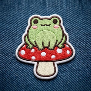 May include: An embroidered patch featuring a light green frog with dark eyes and pink cheeks sitting atop a red mushroom with white spots. The mushroom has a cream-colored stem and a dark brown border, all outlined in white thread.