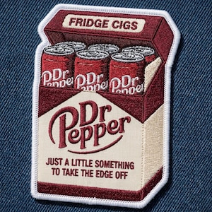 May include: An embroidered patch shaped like a cigarette pack, featuring a maroon and cream color scheme. It displays the text FRIDGE CIGS at the top, Dr Pepper on the cans and front, and the phrase JUST A LITTLE SOMETHING TO TAKE THE EDGE OFF.