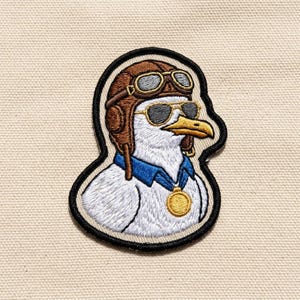 May include: An embroidered patch featuring a white seagull wearing a brown leather aviator cap with goggles, dark sunglasses, a blue collar, and a gold medal hanging from a blue ribbon. The patch has a thick black border and white stitching.