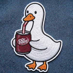 May include: An embroidered patch featuring a white duck with an orange beak and feet. The duck is holding a maroon soda can with the text Dr Pepper and a red straw. The patch has a thick white border and is set against a blue denim fabric background.