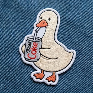 May include: An embroidered patch featuring a light beige duck with an orange beak and feet. The duck is holding a gray soda can with the text Diet Coke in red letters, with a white straw extending from the can to the duck's beak, outlined in white.