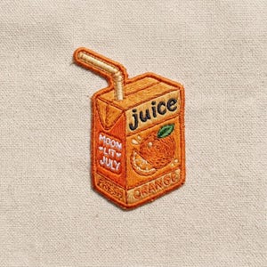May include: An embroidered patch shaped like an orange juice box with a straw. The box is orange with the text juice, moon lit july, and orange embroidered on it. A graphic of an orange slice is centered on the front of the juice box design.