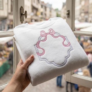 Puede incluir: Sudadera blanca con un diseño de lazo y cinta rosa bordado, enmarcado por un borde plateado. La sudadera doblada se muestra frente a una ventana.