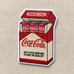 May include: An embroidered patch shaped like a cigarette pack with a red and cream color scheme. The top flap reads FRIDGE CIGS. Inside are red soda cans labeled Coca-Cola. The front features the Coca-Cola logo and the text JUST A LITTLE SOMETHING TO TAKE THE EDGE OFF.