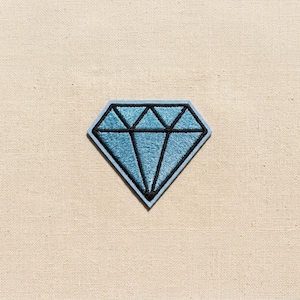 May include: A diamond-shaped embroidered patch featuring light blue thread fill and bold black thread outlines that define the geometric facets of the gemstone design. The patch has a clean, finished edge and a flat, textured surface construction.