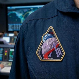 May include: Close-up of a dark blue uniform with a triangular patch. The patch features a purple moon, a blue and white planet, and the names "WISEMAN", "GLOVER", "KOCH HANSEN". The patch has a gold border and red and purple accents.