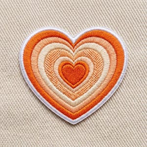 May include: A heart-shaped embroidered patch featuring concentric layers of color in shades of deep orange, light peach, and cream. The design is outlined with a white border and displays a textured, stitched surface throughout the entire shape.