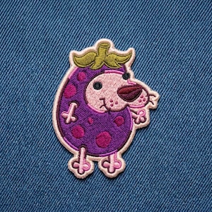 May include: An embroidered patch featuring a cartoon character with a light pink face and a purple, polka-dotted body shaped like a fruit. The character has a green stem on top, small pink limbs, and a dark brown nose, all outlined in light pink thread.