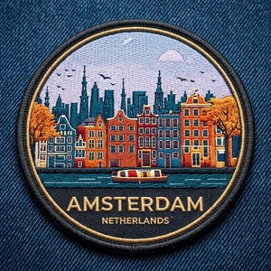 May include: A circular embroidered patch featuring a colorful cityscape of Amsterdam, Netherlands. The design shows orange and blue canal houses, a small boat on the water, and trees with orange leaves. The text AMSTERDAM NETHERLANDS is stitched.