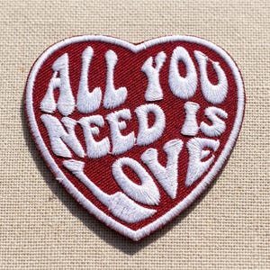 May include: A heart-shaped embroidered patch with a deep red background and a white border. The text ALL YOU NEED IS LOVE is stitched in white thread across the center in a bold, rounded font, filling the entire surface of the heart shape.