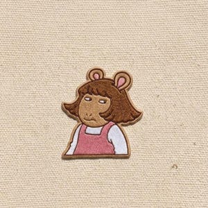 May include: An embroidered patch featuring a brown cartoon character with a bob haircut, wearing a white long-sleeved shirt under a pink pinafore dress. The character has a neutral expression with visible ears and a dark brown outline.