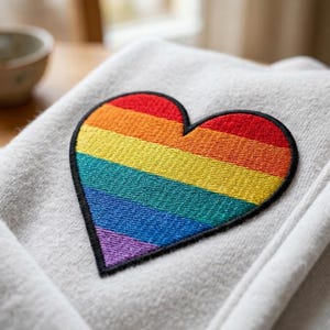 May include: A close-up of a light gray fabric with a large, embroidered heart. The heart features a rainbow of colors: red, orange, yellow, green, blue, and purple, outlined in black. The fabric appears soft and textured.