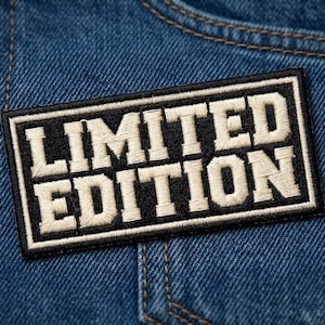 May include: A rectangular embroidered patch featuring the text LIMITED EDITION in bold, off-white capital letters against a solid black background, framed by a thin off-white border, attached to a piece of blue denim fabric with visible stitching.