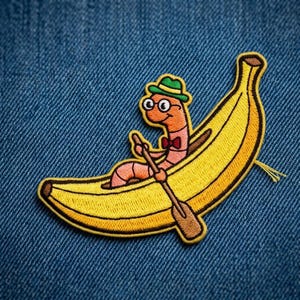 May include: An embroidered patch featuring a light orange worm wearing a green hat, round glasses, and a red bow tie, sitting inside a yellow banana boat and holding a brown wooden oar. The design is stitched with a black border outline.