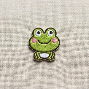 May include: An embroidered iron-on patch featuring a cartoon frog with bright green skin, large white eyes with black pupils, pink circular cheeks, a curved mouth, and a white belly, all outlined in a dark brown thread on a textured fabric.
