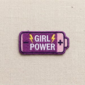 May include: A rectangular embroidered patch shaped like a battery, featuring a purple background with the white text GIRL POWER. The design includes two yellow lightning bolts, a black minus sign on the left, and a black plus sign on the right.