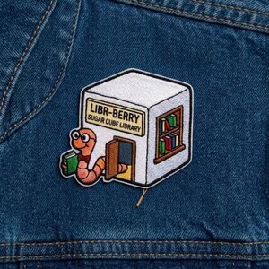 May include: An embroidered patch featuring a white cube-shaped library building with a sign reading LIBR-BERRY SUGAR CUBE LIBRARY. A pink worm wearing glasses and a red bowtie holds a green book while emerging from the open brown door.