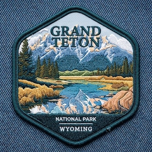 May include: A hexagonal embroidered patch featuring a landscape of the Grand Teton mountains, a river, and pine trees. The text reads GRAND TETON, NATIONAL PARK, and WYOMING. The patch has a dark teal border and is set against a blue denim fabric background.