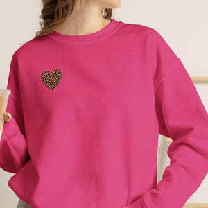 Peut inclure: Un sweat-shirt à manches longues rose vif avec un petit cœur imprimé léopard sur la poitrine gauche. Le vêtement présente une encolure ronde et des poignets côtelés, associé à un short en jean gris et un gobelet en plastique transparent.