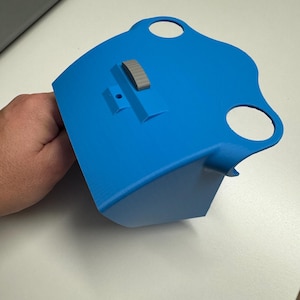 Peut inclure: Un objet bleu vif, imprimé en 3D, avec une partie supérieure incurvée et une base rectangulaire. Il comporte deux ouvertures circulaires et un petit composant rectangulaire gris. L'objet est tenu par une main.