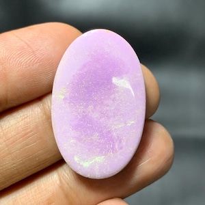 May include: Oval-shaped, light purple gemstone with a polished surface. The stone has subtle white and pale green inclusions, adding texture. The gemstone is held in a hand, showcasing its size and colour.