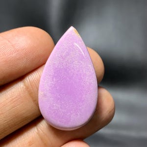 May include: A polished, teardrop-shaped gemstone in a soft lavender colour. The stone has a smooth, glossy surface with subtle white inclusions, held in a hand against a dark background. The gemstone's shape is elongated, with a rounded base and a pointed tip.