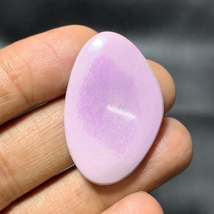 May include: A polished, oval gemstone in a soft lavender colour. The stone has a smooth, glossy surface with subtle colour variations, and is held in a hand against a dark background. The gemstone's shape is reminiscent of a teardrop.