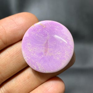May include: A circular, polished gemstone in a light purple shade. The stone has a smooth surface with slight colour and texture variations, and a few golden inclusions. The gemstone is held in a hand against a dark background.