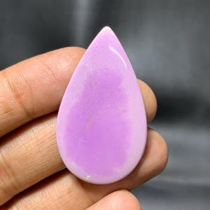 May include: A teardrop-shaped, polished gemstone in a soft lavender hue. The stone has a smooth, glossy surface and a subtle, pearlescent sheen. The gemstone is held in a hand, with a dark background.