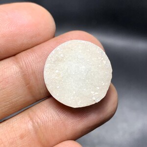 May include: A round, off-white stone with a rough, textured surface. The stone is held in a hand against a dark background. The stone appears to be a natural mineral specimen.