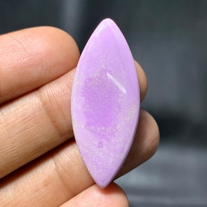 May include: A polished, elongated gemstone in a soft lavender hue. The stone has a smooth, glossy surface with subtle white inclusions, and is held in a hand against a dark background. The shape is reminiscent of a leaf or an eye.