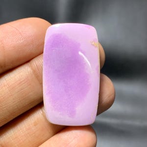 May include: A polished, rectangular gemstone with rounded edges. The stone has a soft lavender hue with lighter, swirling patterns. Small gold-coloured inclusions are visible on the surface. The gemstone is held in a hand.